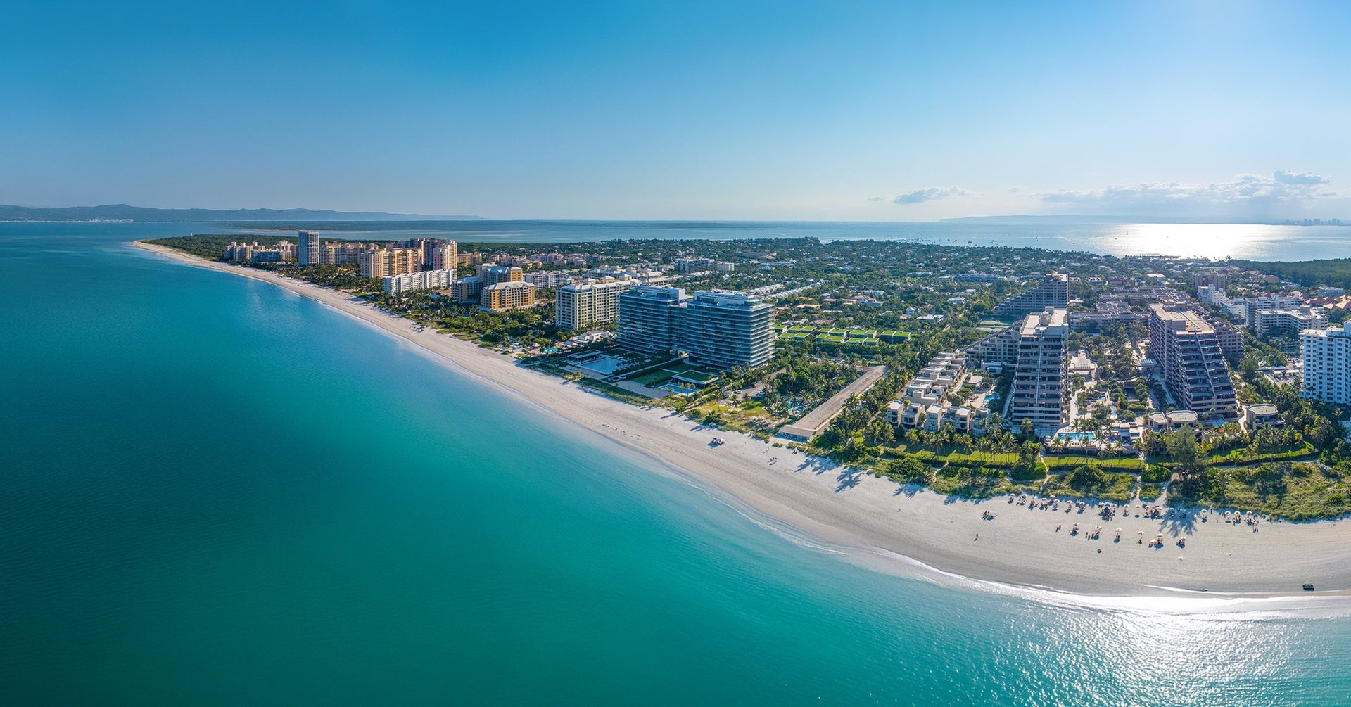 Key Biscayne luxury and ultra luxury condos in an aerial beachfront view with a curved sandy shoreline, turquoise water, oceanfront residences, and a lush island neighborhood stretching to the horizon.