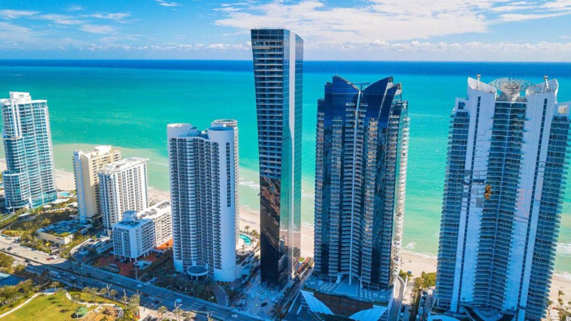 Aerial beachfront view of Muse Residences in Sunny Isles Beach beside turquoise water and neighboring towers, highlighting luxury and ultra luxury condos along the oceanfront.