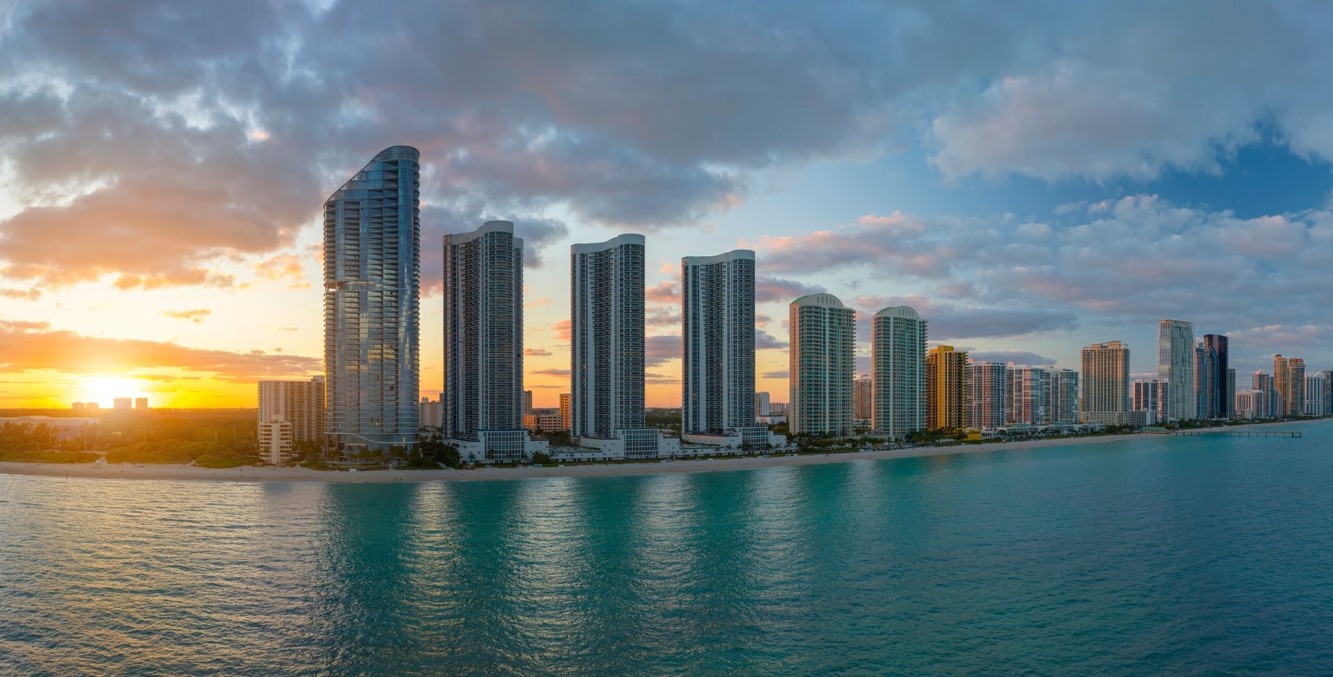 Sunny Isles luxury and ultra luxury condos in a sunset beachfront skyline view with tall glass towers along the shore, dramatic clouds, and calm ocean water reflecting the evening light.